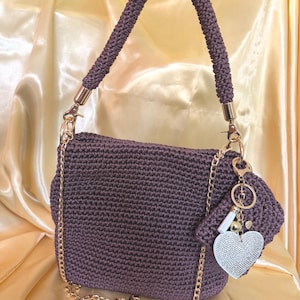 Puede incluir: Un bolso de mano de ganchillo de color morado oscuro con un asa superior gruesa a juego y una correa de hombro de cadena metálica dorada. El bolso está adornado con un llavero dorado con un dije en forma de corazón blanco con cristales.