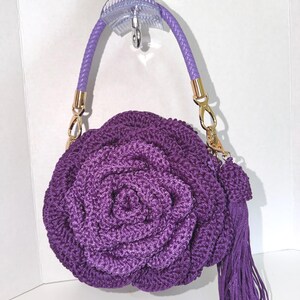 Puede incluir: Un bolso de ganchillo de color morado oscuro con forma de rosa grande en flor. El bolso cuenta con un asa trenzada de color morado claro sujeta con herrajes metálicos dorados y una borla larga a juego que cuelga del lateral del cuerpo.
