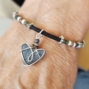 May include: A black elastic bracelet with white and green beads and a silver charm in the shape of a heart. The heart charm is made of a gray stone wrapped in silver wire.