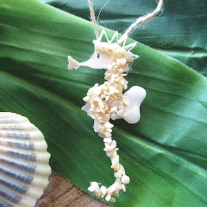 May include: A white seahorse ornament made from seashells, hanging from a twine string. The seahorse is on a green leaf background.