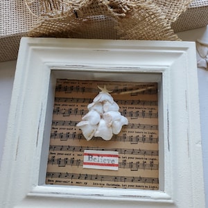 May include: A white framed shadow box with a seashell Christmas tree inside. The tree is made of white seashells and is surrounded by sheet music with the words "Let your heart be light" and "Have yourself a merry little Christmas". There is a red and white fabric banner with the word "Believe" on it.