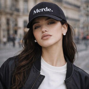 May include: Black baseball cap with the word "Merde" in white lettering. The cap is worn by a person wearing a black bomber jacket and a white crew neck shirt. The person is also wearing diamond stud earrings.