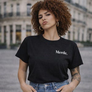 Pode incluir: T-shirt preto com gola redonda com a palavra "Merde" impressa em branco. A camisa é feita de um material macio. A pessoa está usando brincos de argola dourados e jeans azul.