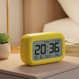Puede incluir: Un reloj despertador digital amarillo que muestra la hora 20:36, con la fecha y el día de la semana. El reloj está sobre una mesita de noche, junto a una pequeña planta y libros. La fecha en el reloj es 13.8. SUN 26.