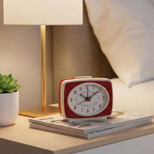Puede incluir: Un reloj despertador retro rojo y blanco se encuentra sobre una mesita de noche, junto a una lámpara y una pequeña planta suculenta en maceta. El reloj tiene una esfera blanca con números y manecillas negras. La mesita es de madera y tiene un cajón blanco.