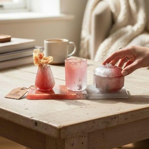 May include: A collection of decorative resin items in shades of pink and red, including a small vase with dried flowers, a tumbler, and a lidded jar. The items are displayed on a wooden table, with a tag that reads "ALBERTS".