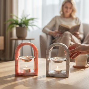 May include: Two decorative candle holders with lit candles. One is pink and the other is gray, both with an arched design. Each has a small dish above the candle. A person is reading a book in the background.