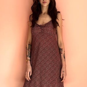 May include: A burgundy slip dress with a small pink polka dot pattern. The dress has a ruffled hem and spaghetti straps. The person is wearing black platform Mary Jane shoes. The background is a solid peach color.