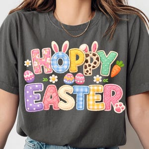 Può includere: T-shirt grigio scuro con una grafica colorata "Hoppy Easter". Il testo presenta vari motivi, tra cui quadretti, floreali e pois, con orecchie di coniglio e una carota. Dettagli di uova di Pasqua e impronte di zampe completano il design.