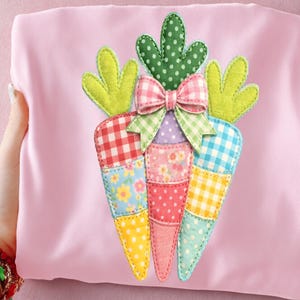 May include: A pink fabric item featuring a colorful, patchwork carrot applique. The carrot has green leaves, a gingham bow, and sections of floral, polka dot, and gingham patterns in various colors. The item is held by a hand.