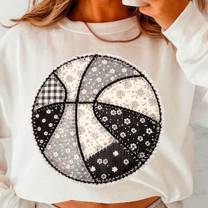 May include: White sweatshirt with a basketball design. The basketball is made from patches of black, white, and grey fabrics, including floral and gingham patterns. The design is stitched onto the sweatshirt.