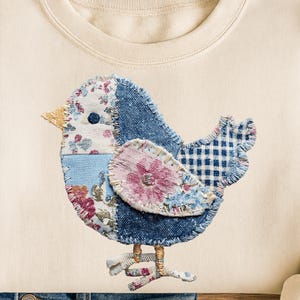 May include: Cream-colored sweatshirt featuring a patchwork bird design. The bird is made of denim, floral, and gingham fabrics, with a yellow beak and delicate stitched details. The bird's body includes a pink flower.