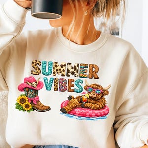 May include: Cream-colored sweatshirt with a colorful graphic that reads "SUMMER VIBES." The graphic features a cowboy boot with a pink hat, sunflowers, and a Highland cow relaxing on a pink inflatable ring.
