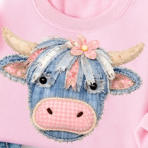 May include: Pink sweatshirt featuring a denim cow face applique. The cow has fabric horns, ears, and a pink gingham nose. The cow's hair is made of blue and white fabric strips, and a pink flower sits on its head.