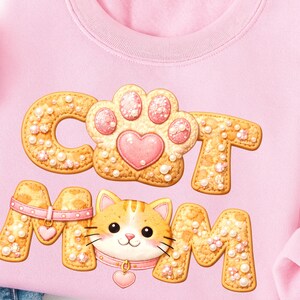 May include: A pale pink sweatshirt featuring the words "CAT MOM" in a cookie-style design. The paw print and cat face are central to the design. The cat has orange and white fur and wears a pink collar. The sweatshirt is a comfortable fit.
