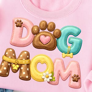 May include: Pink sweatshirt featuring the words "DOG MOM" in a colourful, stitched design. The letters are in various colours and styles, including a paw print, a bone, and a heart.