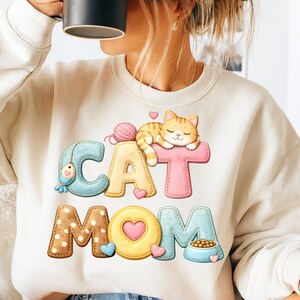 May include: A cream-coloured sweatshirt featuring the text "CAT MOM" in a colourful, whimsical design. The design includes a cartoon orange tabby cat, a ball of yarn, a fish, hearts, and a bowl of cat food. The letters are in various colours.