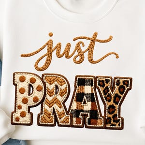 Può includere: Felpa bianca con la scritta ricamata "just PRAY". La parola "just" è in marrone con un carattere corsivo. La parola "PRAY" è in lettere maiuscole con vari motivi, tra cui pois, chevron, quadri e stampa leopardata.