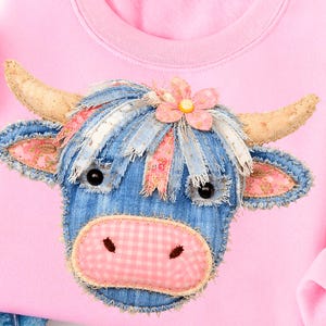 May include: Pink sweatshirt featuring a denim cow face applique. The cow has black eyes, a pink gingham nose, and a flower on its head. The cow's hair is made of blue denim and pink and white ribbons. The horns are tan.