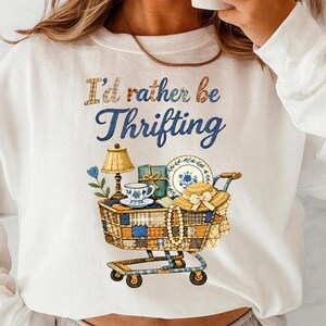 Op de afbeelding: Witte sweatshirt met de tekst "I'd rather be Thrifting". De afbeelding toont een winkelwagentje gevuld met vintage items zoals een lamp, een theekopje en borden, wat een liefde voor kringlopen en vintage vondsten suggereert.