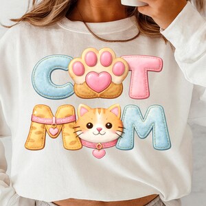 May include: White sweatshirt with the text "CAT MOM" in a colorful, cartoonish design. The "C" and "M" are light blue, the "T" and paw print are pink, and the "O" is a paw print. The "A" and the cat are yellow and orange.