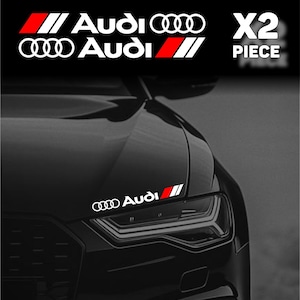 May include: A black car with white and red Audi decals. The image shows the front of the car with the Audi logo and the text "X2 PIECE" in white. The decals are on the bonnet of the car.