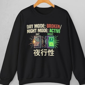 Pode incluir: Camisola preta com um design gráfico com duas ilustrações de interruptores. O texto diz "DAY MODE: BROKEN/ NIGHT MODE: ACTIVE" com caracteres japoneses por baixo. O interruptor do dia está quebrado e o da noite está ligado.