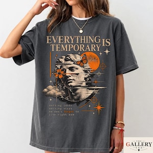 May include: A charcoal gray short-sleeve t-shirt featuring a graphic print of a classical statue head, a butterfly, a sun, and clouds. The text reads "EVERYTHING IS TEMPORARY" and "nothing lasts nothing stays so don't forget to live right now."