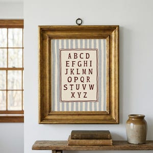 May include: A framed alphabet print with a gold-colored ornate frame. The print features a cream-colored background with vertical blue stripes. The alphabet is displayed in a maroon-colored font. The frame is hanging on a white wall.
