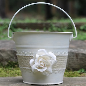 May include: White metal bucket with a handle, decorated with burlap and lace. A white fabric rose is attached to the front.