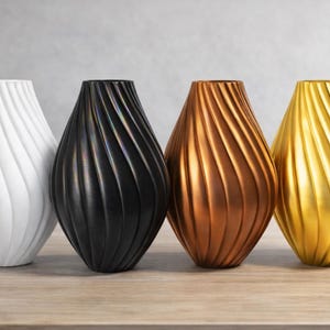 Könnte beinhalten: Vier dekorative Vasen in einer Reihe, jede mit einer einzigartigen Farbe und Design. Die Vasen sind weiß, schwarz, bronze und gold, mit einem strukturierten, wirbelnden Muster. Sie sind tropfenförmig und stehen auf einer Holzoberfläche.