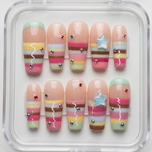 Pode incluir: Um conjunto de dez unhas postiças numa caixa de plástico transparente. As unhas apresentam uma base nude com riscas horizontais multicoloridas, incluindo rosa, amarelo, castanho e verde. Algumas unhas têm uma estrela azul ou pequenas pedras brilhantes.