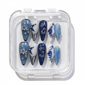 May include: A set of ten false nails in a clear plastic case. The nails feature various designs, including a star and silver accents, a white star on a silver base, and the text "THIS IS FOR" on a navy blue background. Other nails have blue heart and feather designs.