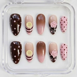 May include: A set of ten artificial nails in a clear plastic case. The nails feature various designs, including brown polka dots, button accents, ombre effects, and floral embellishments. The color palette includes shades of brown, pink, and beige.