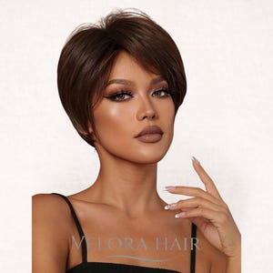 May include: A short, dark brown wig with a side-swept bang. The wig has a natural-looking style and is designed to frame the face. The text "VELORA HAIR" is visible at the bottom of the image.