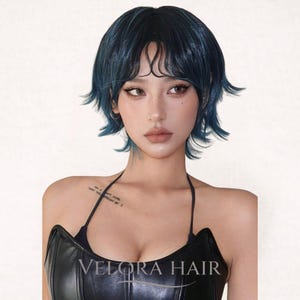 May include: A woman with a short, dark blue wig with bangs. The wig has a layered style. She is wearing a black top with the text "VELORA HAIR".