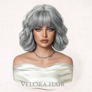 May include: A silver-grey, wavy wig with bangs, displayed on a mannequin bust. The wig has a natural-looking texture and is styled with soft waves. A silver necklace is visible. The text "VELORA HAIR" is printed on the bust.