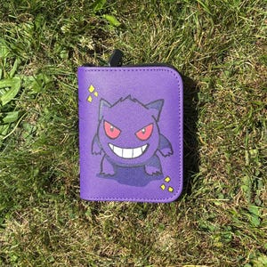 May include: A rectangular purple wallet featuring a graphic of the character Gengar with red eyes and a wide white grin. The wallet has a black zipper closure at the top and is decorated with small yellow diamond-shaped accents on the front surface.