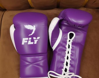 May include: A pair of purple boxing gloves with white accents. The gloves feature the word "FLY" and a graphic of a bird. The gloves are laced up with white laces.