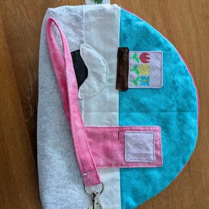 May include: A handmade, quilted pouch with a pink lanyard and a silver clasp. The pouch features a patchwork design with white, light blue, and turquoise fabrics. A small charm of a camper hangs from the zipper. The pouch has a small tag with a mountain scene.