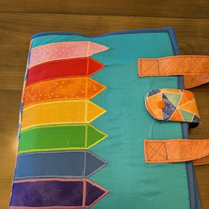 May include: A quilted, teal-colored book cover with a rainbow crayon design. The crayons are pink, red, orange, yellow, green, blue, and purple. It has orange straps and a button closure. The edges are trimmed with blue.