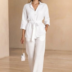 Può includere: Una camicia bianca abbottonata con maniche a tre quarti e una cintura da annodare in vita, abbinata a pantaloni a gamba larga nello stesso tessuto bianco. Una piccola borsa bianca con tracolla a catena dorata è tenuta di lato all'outfit.