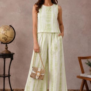 May include: A sleeveless top and wide-leg trousers set featuring a light green and white vertical tie-dye pattern. The outfit is paired with a small rectangular woven handbag that has a white flap and a prominent gold circular buckle detail.