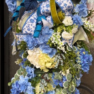 May include: A floral wreath featuring blue hydrangeas, white flowers, and green foliage. The wreath is adorned with blue and white patterned ribbons, teal ribbons, and a touch of olive green velvet ribbon. A decorative accent for doors or walls.