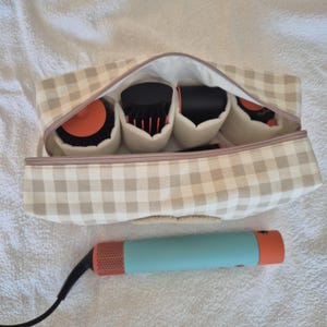 May include: A beige and white gingham patterned cosmetic bag with a zipper, containing hair styling tools. The bag holds several black and orange hair styling attachments. A teal and orange hair styling tool with a black cord is below the bag.