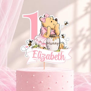 May include: A cake topper featuring a large pink number one, a cartoon bear wearing a pink dress and bow, and a honey pot labeled Hunny. A white banner at the bottom displays the name Elizabeth in pink script, surrounded by small flying bees.