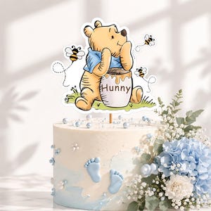 Può includere: Un topper per torta raffigurante un orso giallo con una maglietta blu, seduto accanto a un barattolo di miele con l'etichetta Hunny, circondato da tre piccole api. Il topper è posizionato su una torta bianca decorata con impronte.