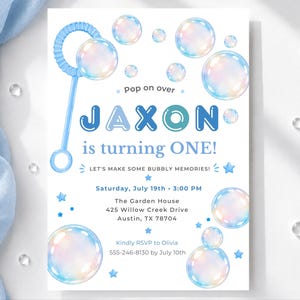 Può includere: Un biglietto d'invito per compleanno con una bacchetta per bolle blu e bolle iridescenti. Il testo recita: pop on over JAXON is turning ONE! LET'S MAKE SOME BUBBLY MEMORIES! Saturday, July 19th, 3:00 PM, The Garden House, 425 Willow Creek Drive, Austin, TX 78704, Kindly RSVP to Olivia 555-246-8130 by July 10th.