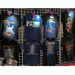May include: A collection of eight black and navy blue short-sleeve t-shirts featuring Eric Clapton graphics. The shirts display various designs including guitars, concert tour dates for the 2026 American Tour, and the text Eric Clapton Slowhand.