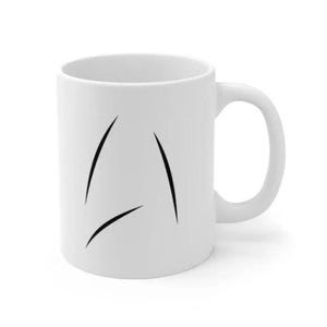 Pode incluir: Caneca de cerâmica branca com uma alça curva. A caneca apresenta três linhas pretas e curvas na frente. A caneca é projetada para bebidas quentes.